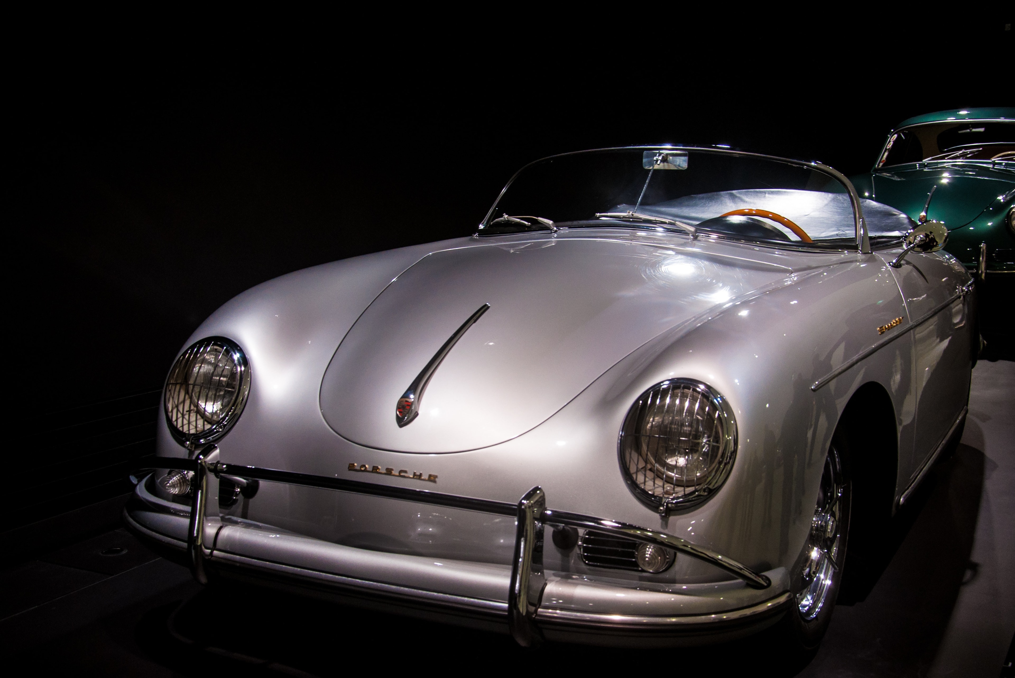 Porsche 356 Speedster gris argent, présentée sous éclairage d’exposition, un coupé vert à l’arrière-plan